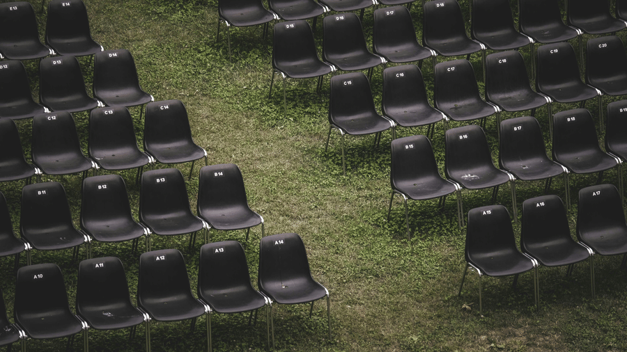 Rows of empty, dark plastic chairs set up in an open-air venue on a slightly overgrown green grass lawn.