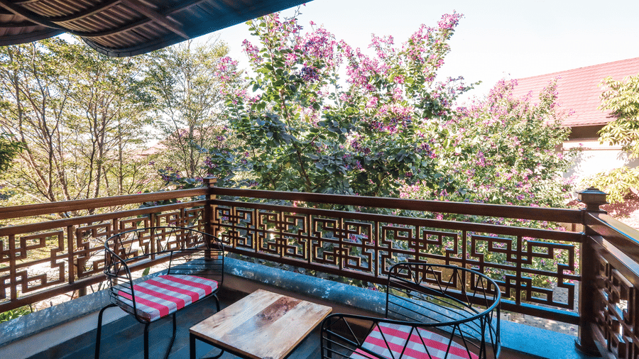 A balcony shows chairs with a table placed beside a railing overlooking trees and nearby buildings at Ananta Spa & Resort, Jaipur.