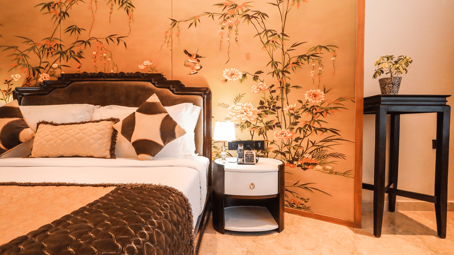 A room shows a bed with cushions, side tables on both sides, plants placed beside the tables and a wall panel behind the bed at Ananta Spa & Resort, Jaipur.