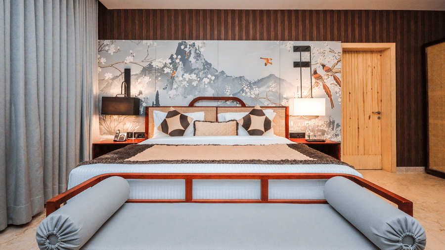 4. Bed Frame A room shows a bed with a large cushioned frame, side tables, artwork above the bed and curtains on a window at Ananta Spa & Resort, Jaipur.