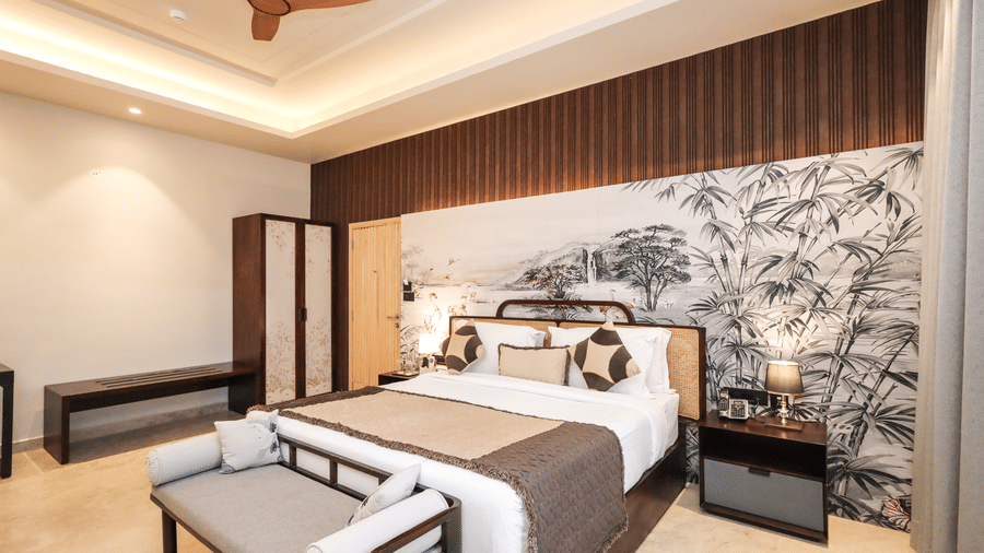 An interior shows a bed with side tables, a bench at the foot of the bed, a wall artwork behind the bed and storage units on the side at Ananta Spa & Resort, Jaipur.