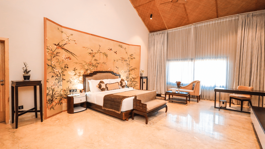 A room shows a bed with side tables, a bench at the foot of the bed, a seating area with chairs and a large window with curtains at Ananta Spa & Resort, Jaipur.