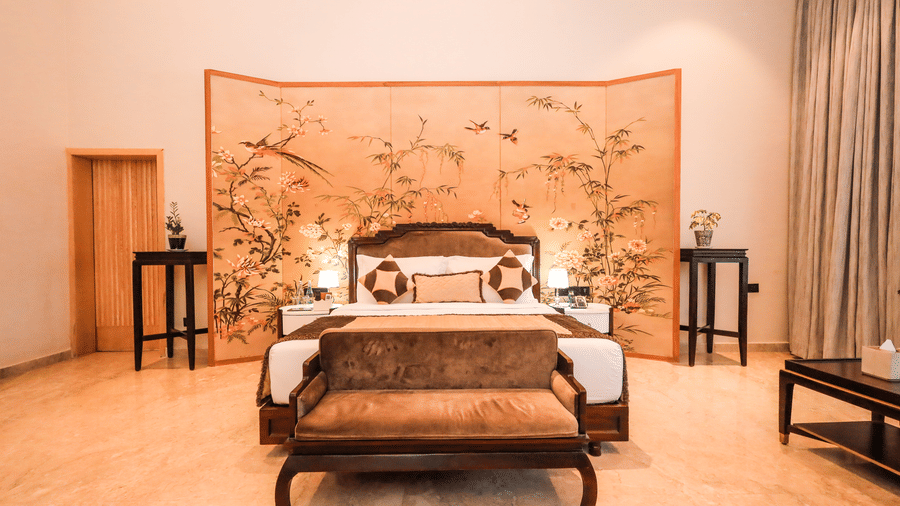 A room shows a bed with side tables, plants on both sides, a bench at the foot of the bed and a wall panel behind the bed at Ananta Spa & Resort, Jaipur.