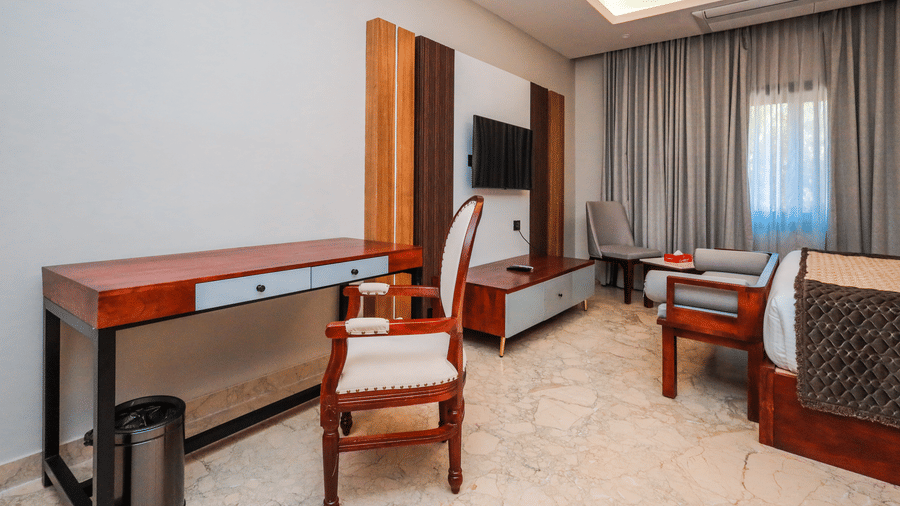 A room shows a desk with a chair, a television on a wall, a bed with cushions and curtains on a window at Ananta Spa & Resort, Jaipur.