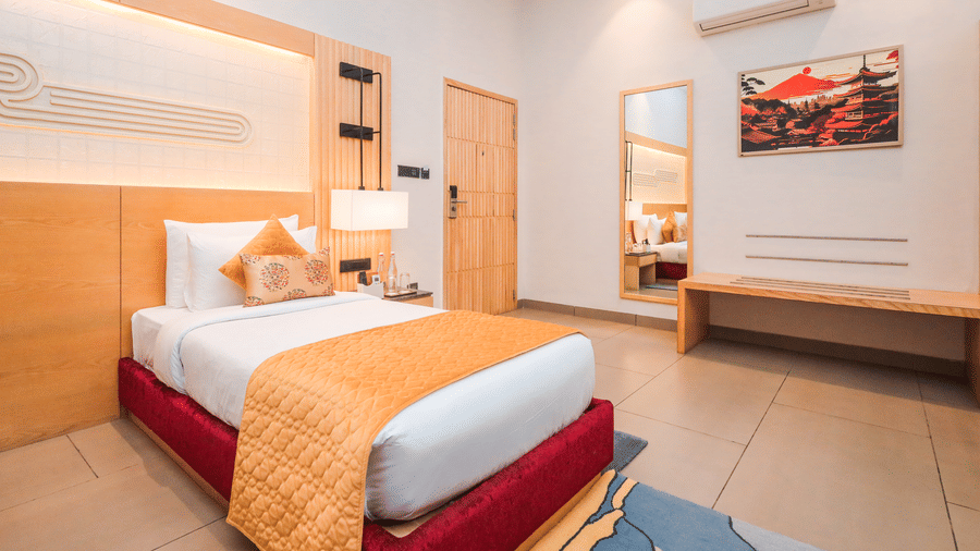 A room shows a bed with cushions, a side table with a lamp, wall art above the bed and a door leading to another area at Ananta Spa & Resort, Jaipur.