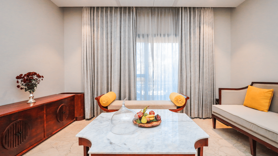 A room shows a bed with cushions, side tables, a seating bench, curtains on a window and a table with fruit at Ananta Spa & Resort, Jaipur.