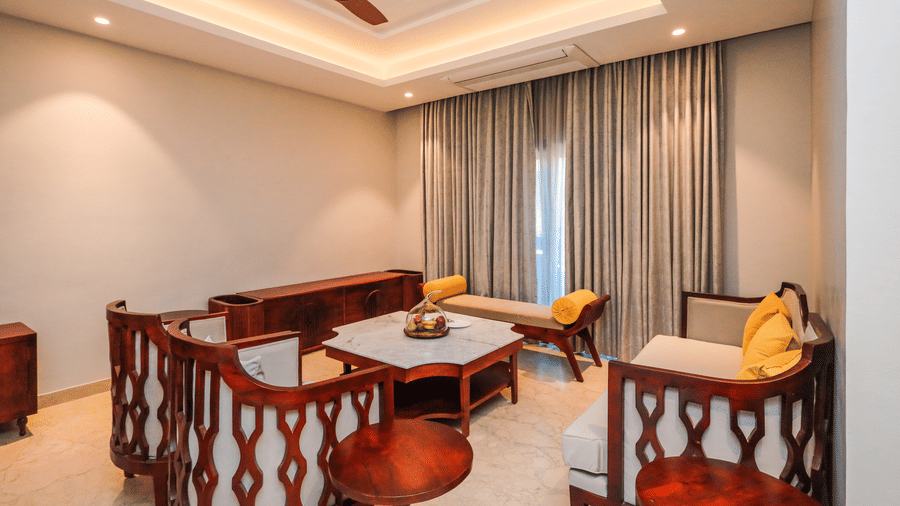 A room shows chairs with a table, long seating with cushions, a window with curtains and a ceiling fan at Ananta Spa & Resort, Jaipur.