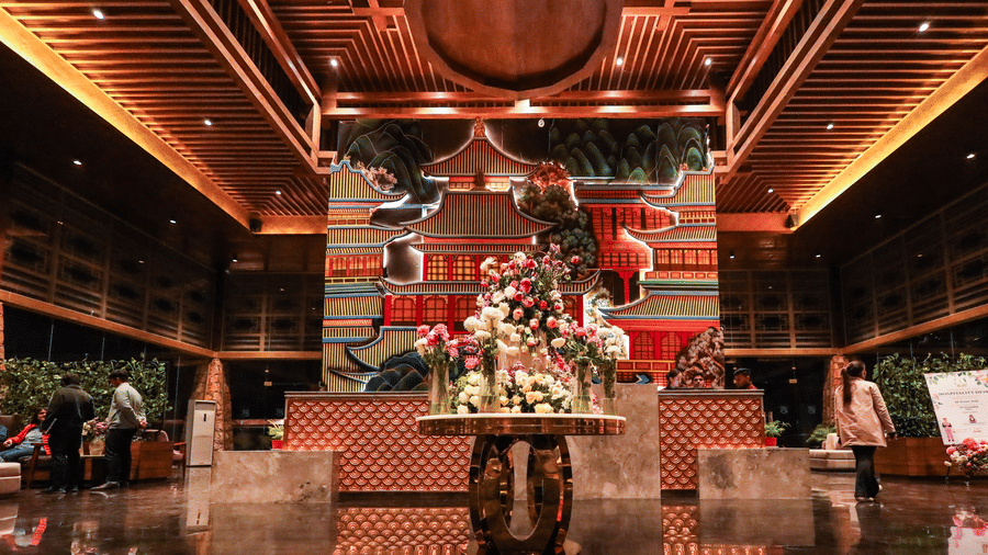 The luxurious lobby at Ananta Spa & Resort, Jaipur, features a round floral arrangement, a vibrant pagoda mural, and wood ceilings.