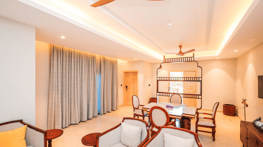 A room shows chairs with a table, a long seating area with cushions, curtains on windows and a ceiling fan at Ananta Spa & Resort, Jaipur.
