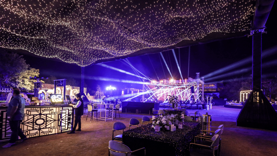 An outdoor event at Ananta Spa & Resort, Jaipur, features a stage with blue lighting and a striking canopy of string lights.