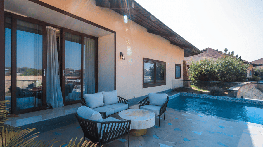 An outdoor area shows a seating arrangement beside a pool, a villa exterior with sliding doors and plants near the water at Ananta Spa & Resort, Jaipur.