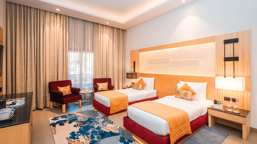 A room with two single beds, seating, lamps and curtains arranged around a main space at Ananta Spa & Resort, Jaipur.