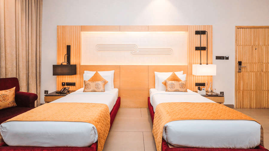 A room shows two beds with side tables, a lamp, a wall panel above the beds and a doorway on the side at Ananta Spa & Resort, Jaipur.