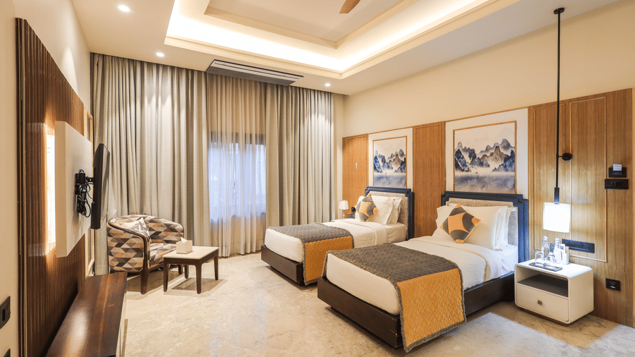 A room shows two beds with side tables, a seating table with chairs, a window with curtains and overhead lighting at Ananta Spa & Resort, Jaipur.