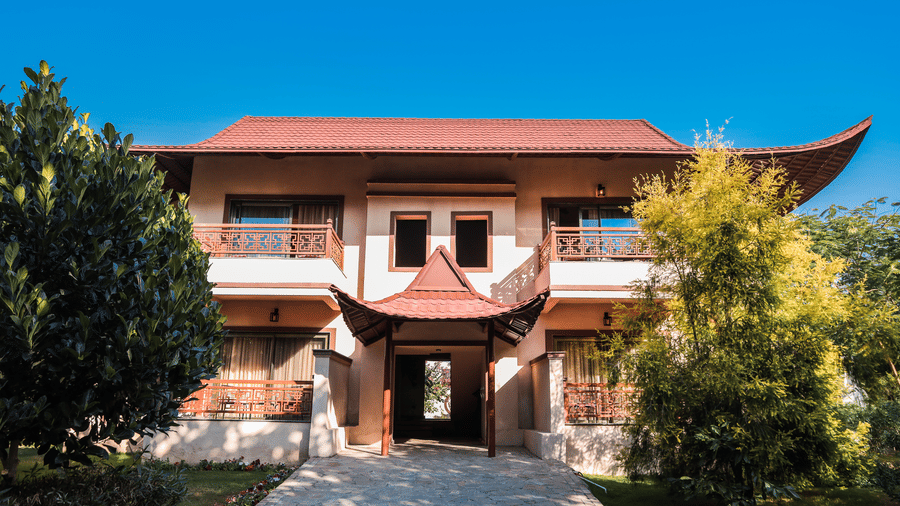 2-storey building with Asian-style red tiled roof, decorative balconies, and a central entrance, surrounded by trees, plants, and a garden under a clear blue sky at Ananta Spa & Resort, Jaipur, one of the finest 5-star Resorts in Jaipur.