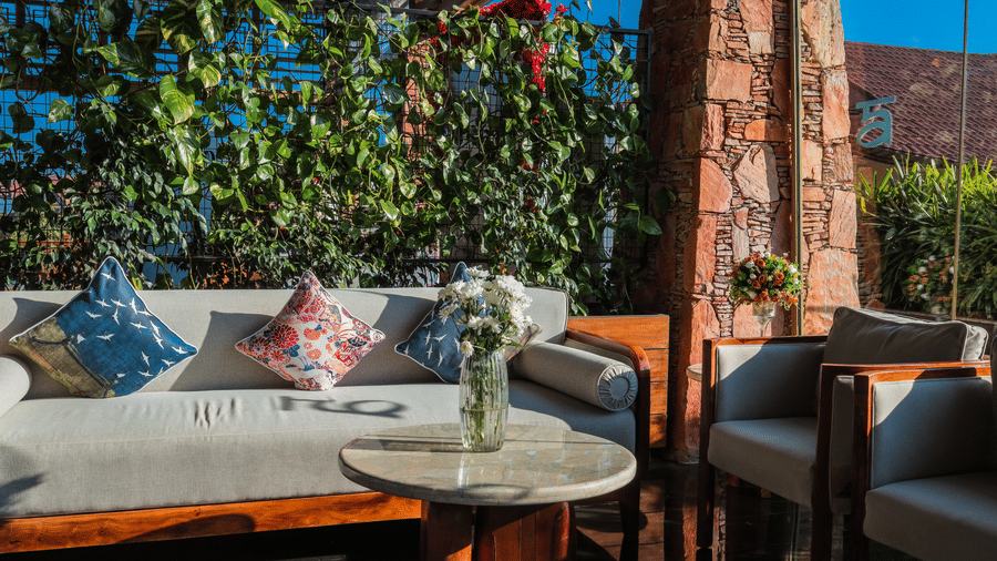 A bright outdoor seating area at Ananta Spa & Resort, Jaipur, features a white sofa with patterned cushions next to a stone column.