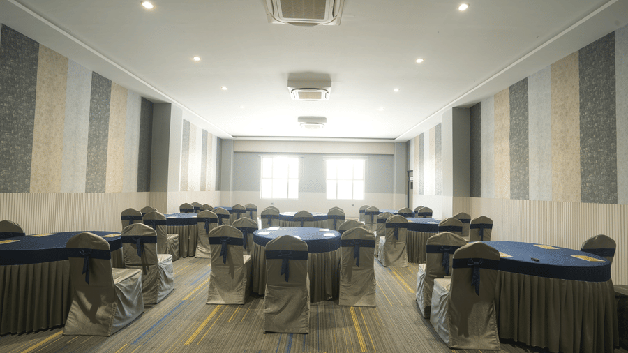 A symmetrical view of a formal banquet hall with round tables draped in blue and silver, set under bright, recessed ceiling lights at Apartel by Aarin, Oragadam.
