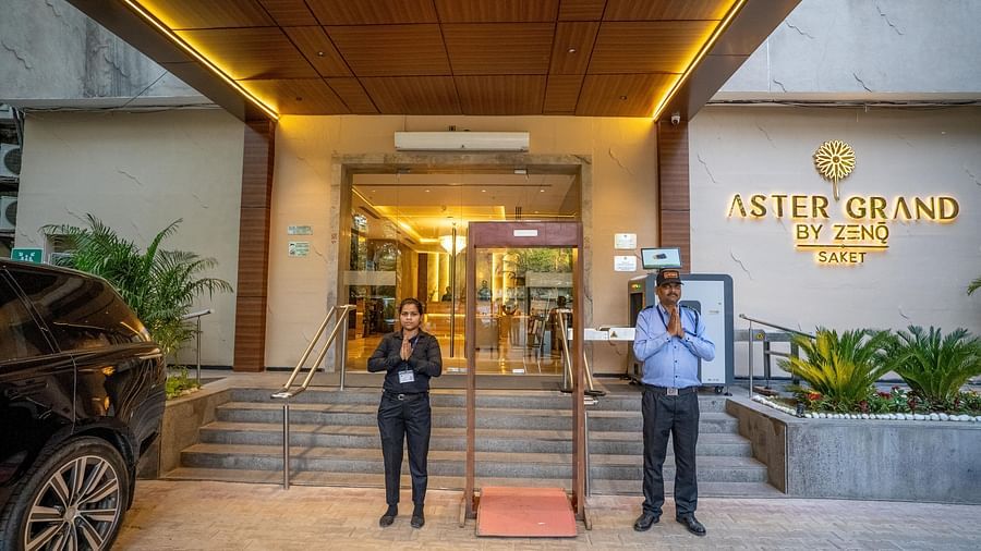 Entrance of ASTER GRAND BY ZENQ SAKET with a car, plants and two staff members standing in the front by joining their hands.