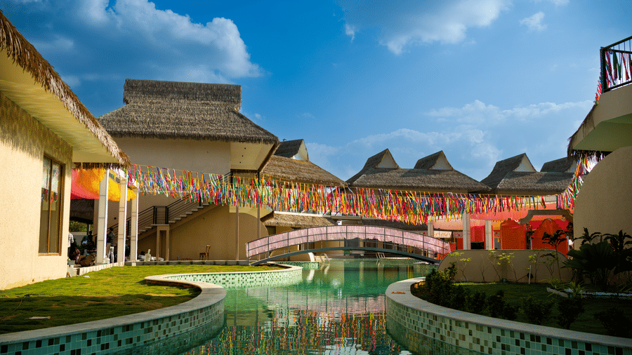 A small bridge crosses a narrow water feature between thatched-roof buildings under a blue sky at Avanya Resorts N Retreat.