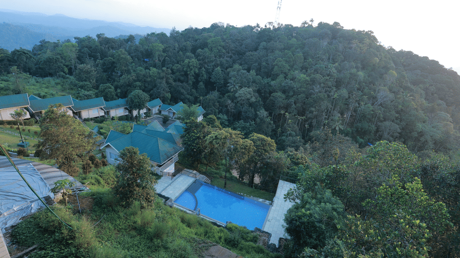 A line up of villas with a turquoise roof along with an outdoor swimming pool surrounded by lush greenery at Aveda Mountains and Mist Resort and Spa, Munnar.