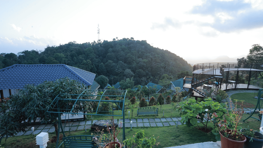 The garden area with a tiled pathway going through it, overlooking a mountain against a bright cloudy sky at Aveda Mountains and Mist Resort and Spa, Munnar.