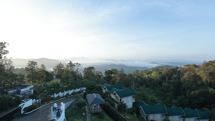 A wide-angle shot of Aveda Mountains and Mist Resort and Spa, Munnar, featuring a line of villas surrounded by lush greenery, with a misty valley backdrop.