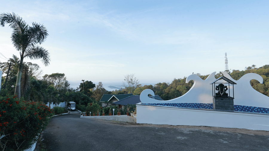 A grey pathway filled with gravel, lined with green bushes, featuring a stone Ganesh idol in front of the driveway at Aveda Mountains and Mist Resort and Spa, Munnar.