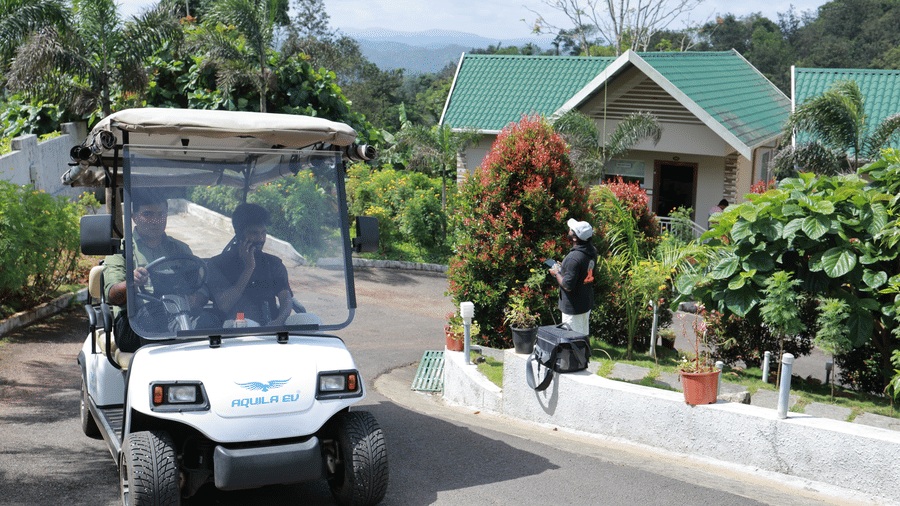 A golf  cart with 2 people sitting in it, driving as they pass through the villas at Aveda Mountains and Mist Resort and Spa, Munnar, on a bright sunny day.