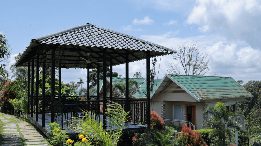 A roofed outdoor seating area besides a tiled pathway at Aveda Mountains and Mist Resort and Spa, Munnar, featuring a clear blue sky with clouds in the background.