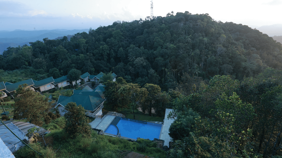 The outdoor swimming pool at Aveda Mountains and Mist Resort and Spa, Munnar, surrounded by small trees and bushes along with a lush green hill in the background.