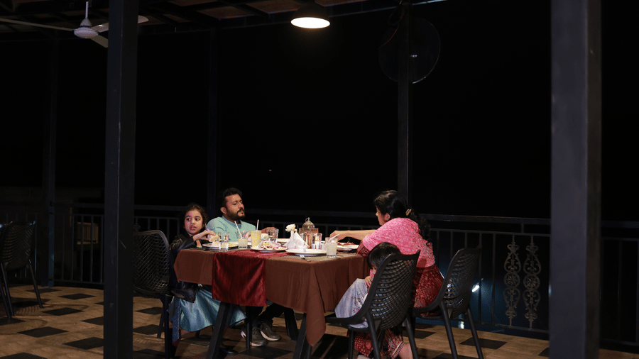 A family of 3 dining at the Machaan restaurant under the dim lighting of a lamp hanging above them at Aveda Mountains and Mist Resort and Spa, Munnar.