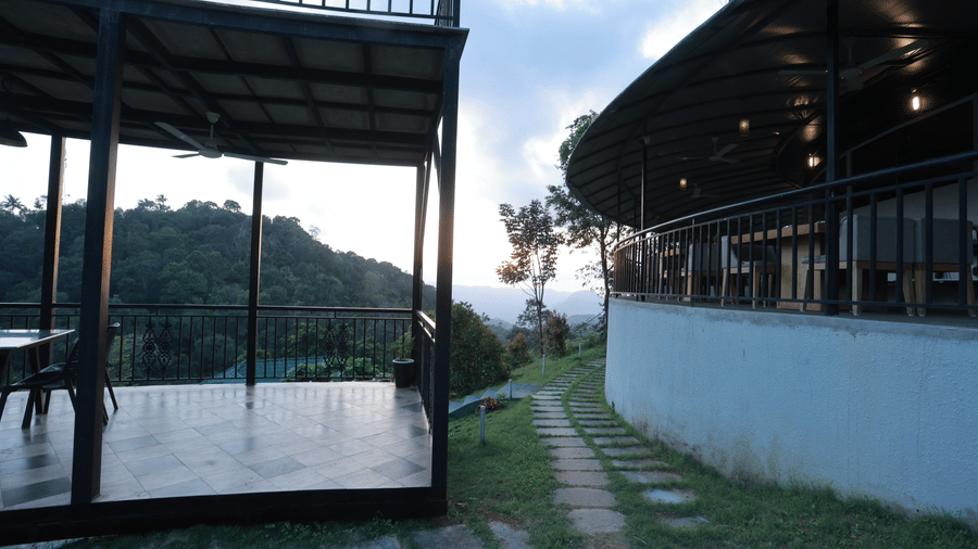 A pathway paved between the outdoor dining area and a wall of a resort through a grassy lawn patch at Aveda Mountains and Mist Resort and Spa, Munnar.