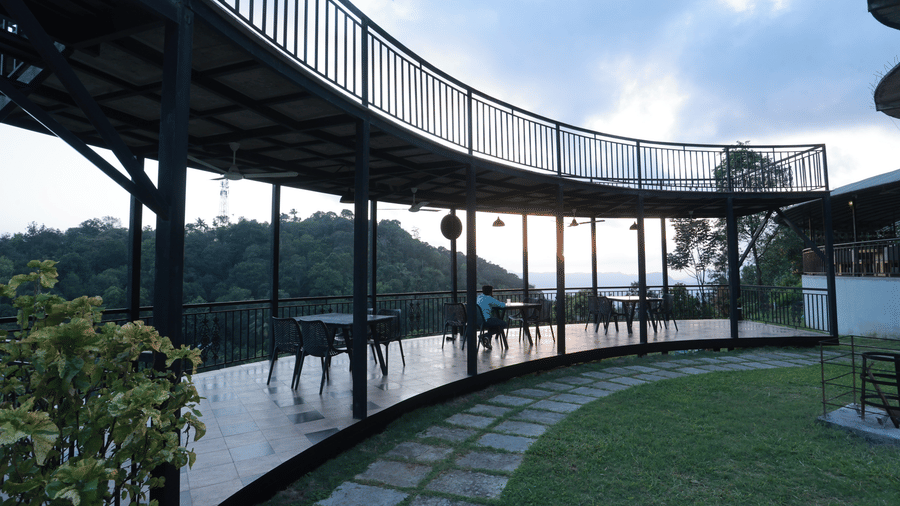 Full view of the outdoor dining area of the Machaan restaurant arching inwards towards the lawn with a tiled pathway at Aveda Mountains and Mist Resort and Spa, Munnar.