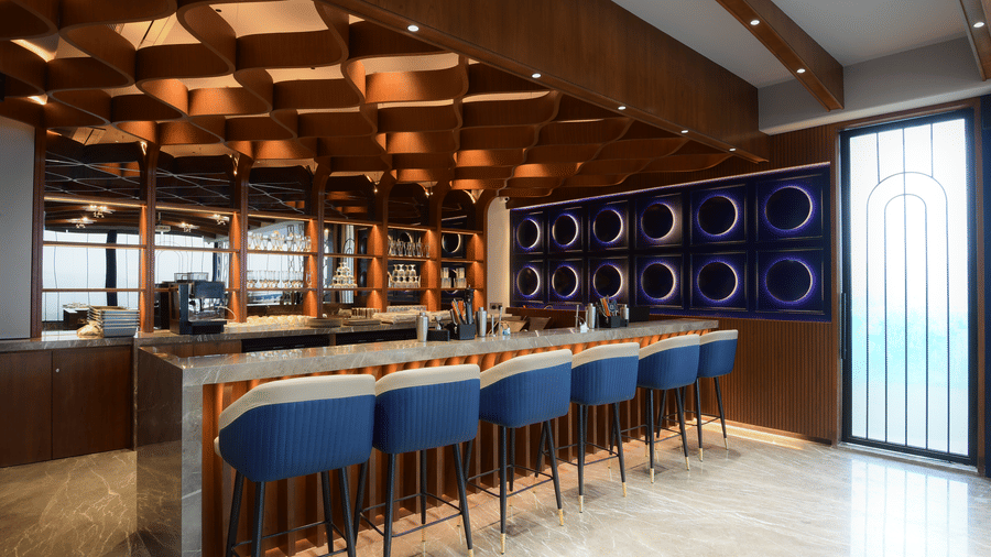 An indoor bar lounge area showcasing a long serving counter, cushioned bar stools, and an intricate wooden ceiling design at Awesome Palace, Guwahati Airport.