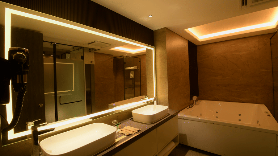 A bathroom vanity counter showing 2 countertop sinks, a large backlit rectangular mirror, and a bathtub at Awesome Palace, Guwahati Airport.