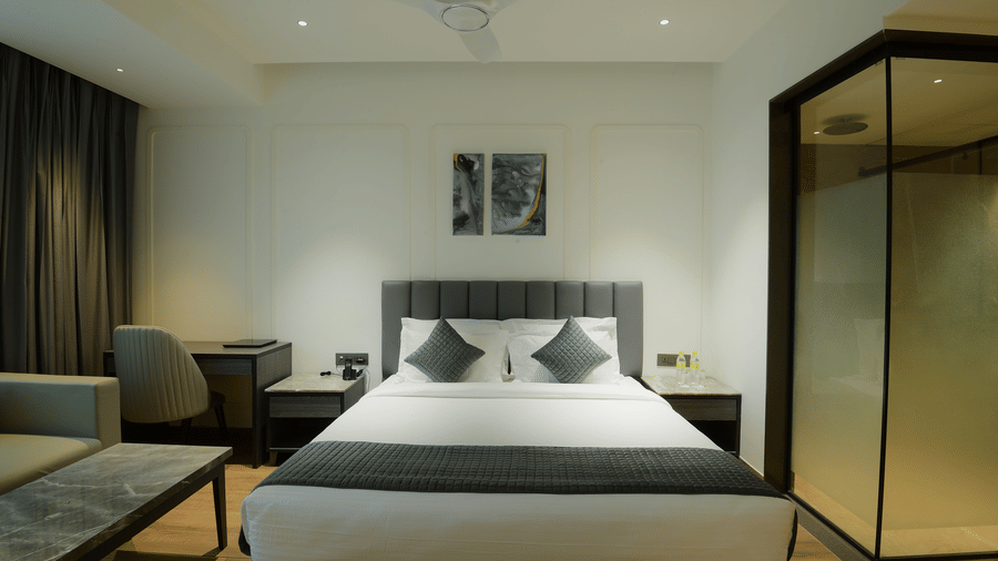 A king size bed flanked by nightstands, a study desk with a chair, and a glass partition at Awesome Palace, Guwahati Airport.