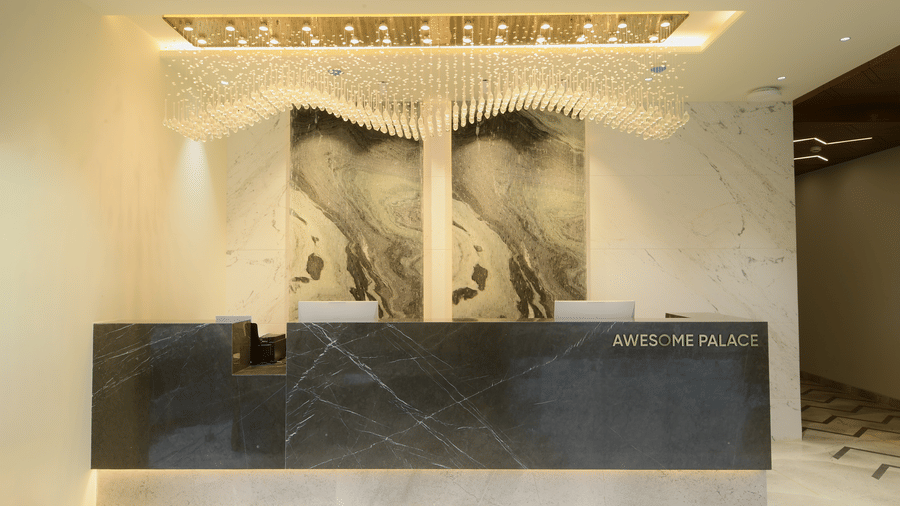 A marble front desk with a textured backdrop panel, computer screens, and decorative hanging lights above at Awesome Palace, Guwahati Airport.
