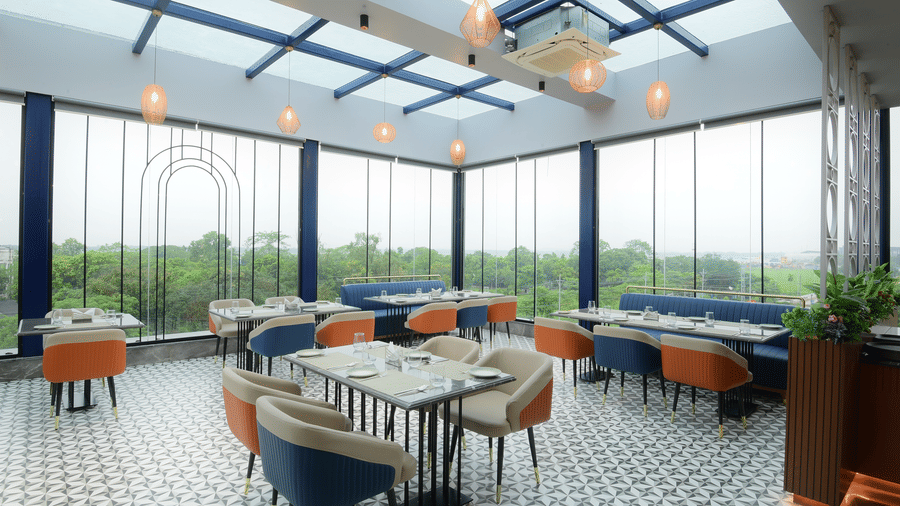 A bright restaurant dining space enclosed by large glass windows, multiple dining tables, and hanging light fixtures at Awesome Palace, Guwahati Airport.