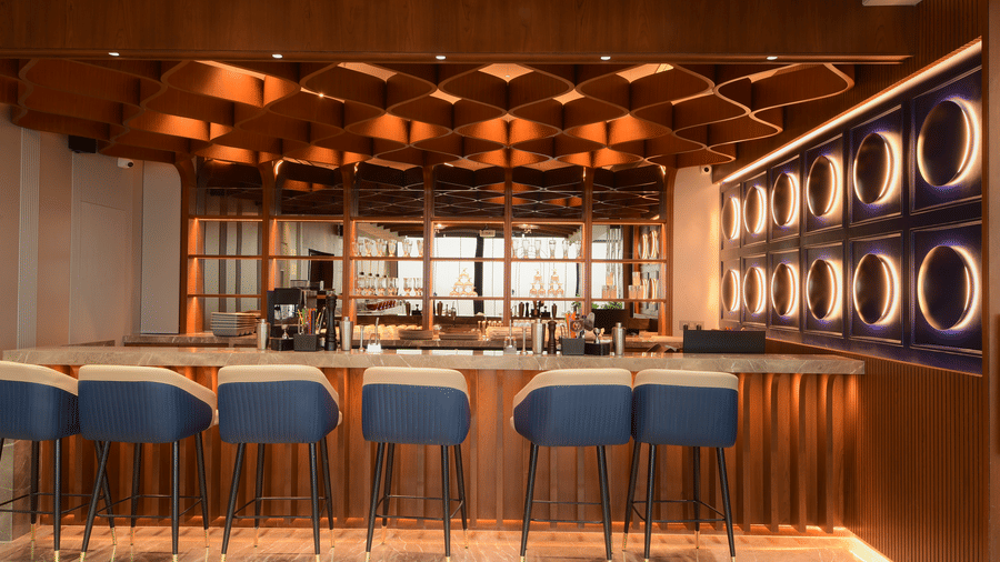A modern bar area featuring cushioned counter stools, a smooth stone countertop, and a decorative wooden ceiling lattice at Awesome Palace, Guwahati Airport.