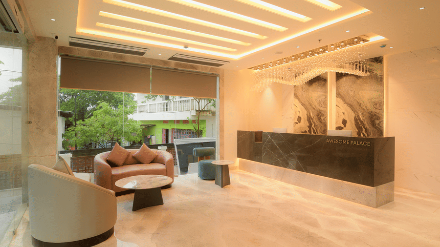 A marble reception desk flanked by a cosy seating area, large windows, and bright linear ceiling lights at Awesome Palace, Guwahati Airport.