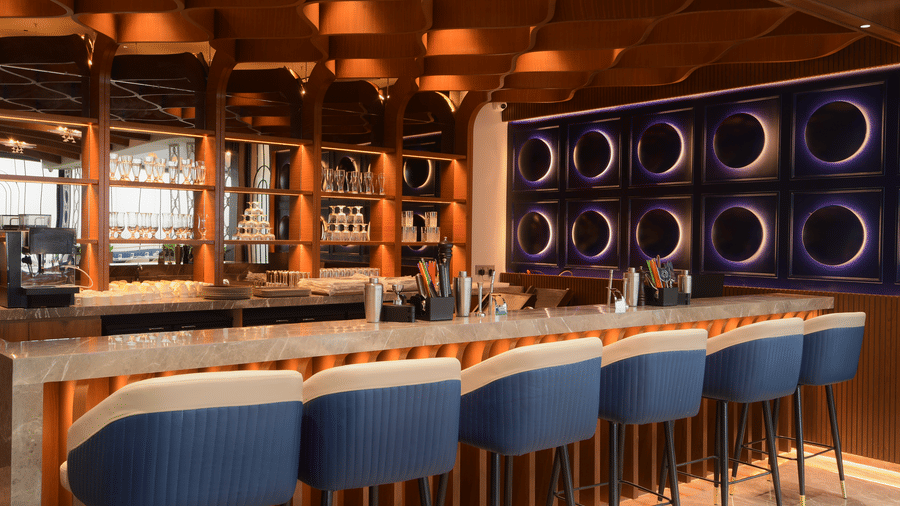 A stylish bar counter setup with a row of high stools, an illuminated under counter, and circular wall light displays at Awesome Palace, Guwahati Airport.