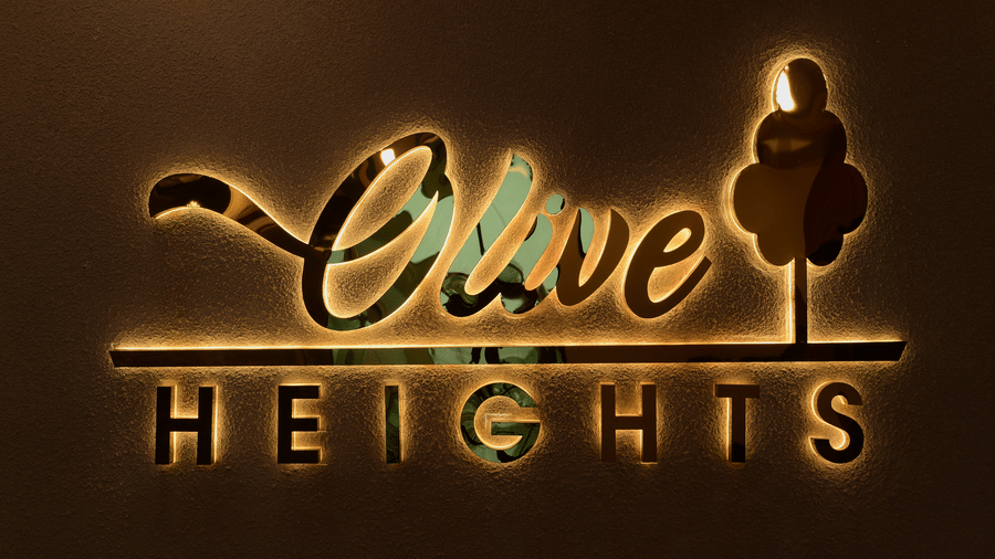 An illuminated wall sign displaying the text Olive Heights, a stylized tree logo, and a dark background surface at Awesome Palace, Guwahati Airport.