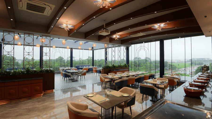 A wide view of a restaurant interior showcasing set dining tables, a buffet service counter, and large glass windows at Awesome Palace, Guwahati Airport.