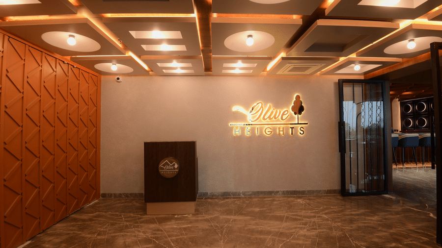 A hotel reception area featuring a dark wooden desk counter, a patterned wall accent, and a glowing brand logo sign at Awesome Palace, Guwahati Airport.