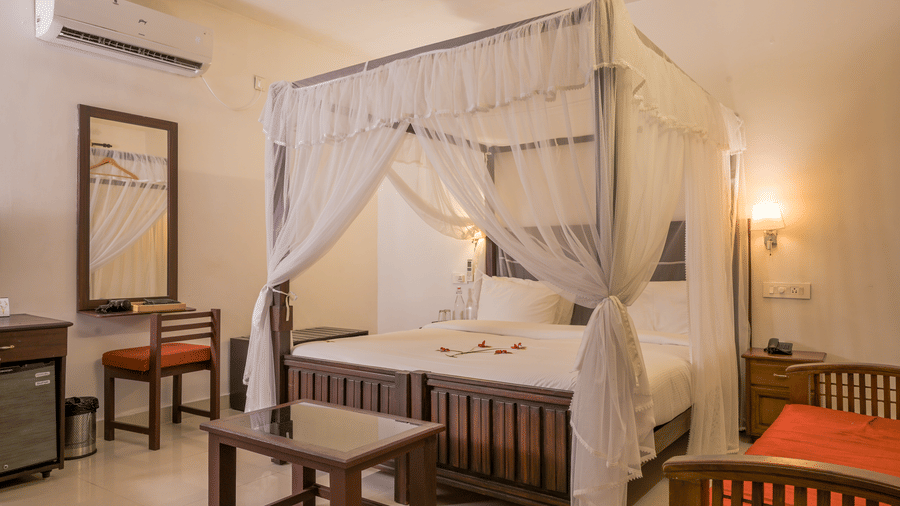 Interior of Ayur Villas at Ayur on the Beach Nattika featuring a four poster canopy bed with white drapes wooden furniture warm wall lighting and a classic traditional decor setting.