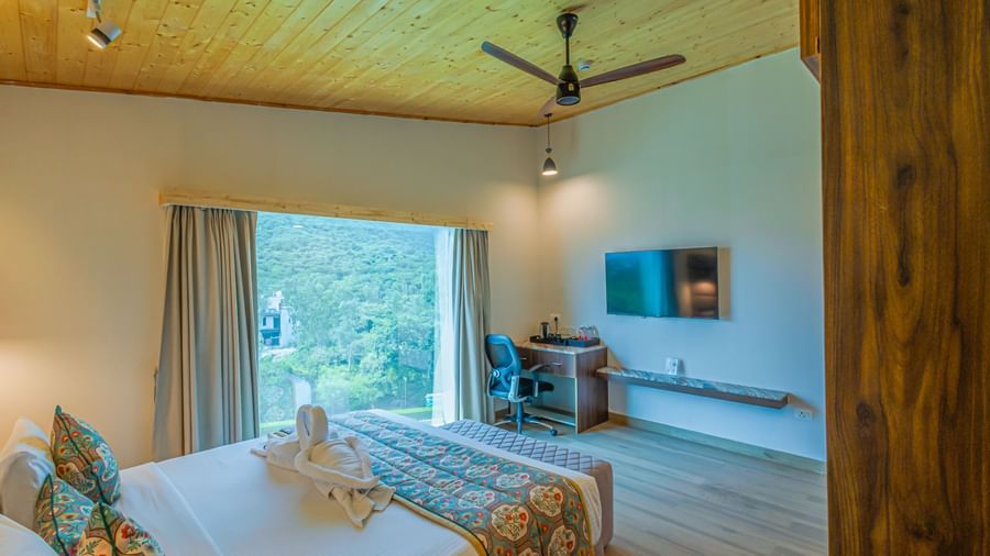 A bright bedroom with a wooden ceiling, fan, and large windows offering a scenic view at Bamboo Saa Resort & Spa, Udaipur.
