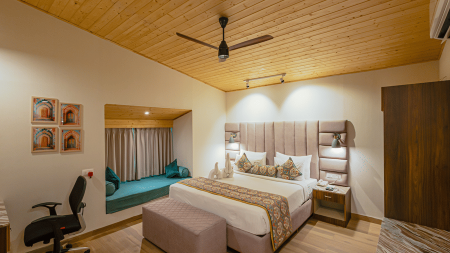 One of the best rooms in Udaipur at Bamboo Saa Resort & Spa with wooden ceiling, double bed with cushions and runner, bench at the foot, bedside tables with lamps, telephone, wardrobe, desk chair, wall art, and window seating area.