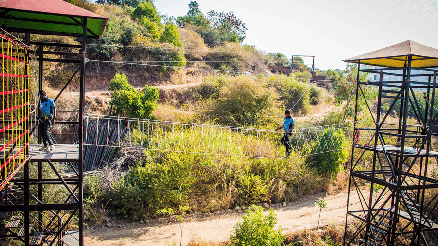 An adventure resort in Udaipur featuring an outdoor setup with a suspended rope bridge between metal platforms, surrounded by greenery and uneven terrain, where two individuals carefully walk across - Bamboo Saa Resort & Spa, Udaipur