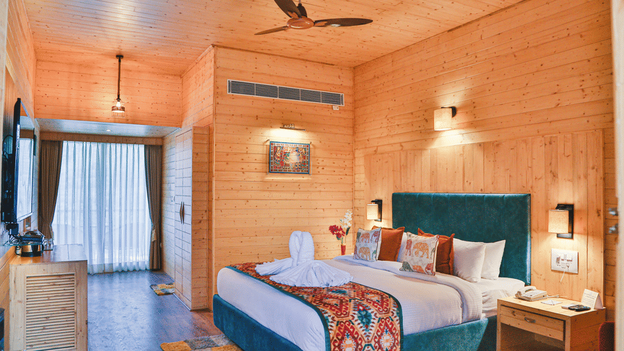 A spacious room at Bamboo Saa Resort & Spa, Udaipur, featuring panelled walls, a plush bed with colourful textiles, bedside tables, a ceiling fan, a wall-mounted television above a wooden console, and a curtained window.