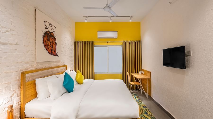 An side angle interior image of the standard room at Bedzzz Varanasi showcases a spacious layout, featuring a comfortable double bed, a cozy floor mat, a stylish study table and chair, a charming coffee table, elegant curtains, an air conditioner for a ref